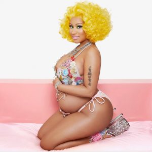 Nicki Minaj in a gold wig and bikini kneeling on a sofa showing the side profile of her pregnant belly.