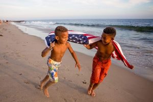black boys running on the beach with the american flag blowing in the wind behind them