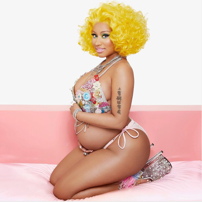 Nicki Minaj in a gold wig and bikini kneeling on a sofa showing the side profile of her pregnant belly.