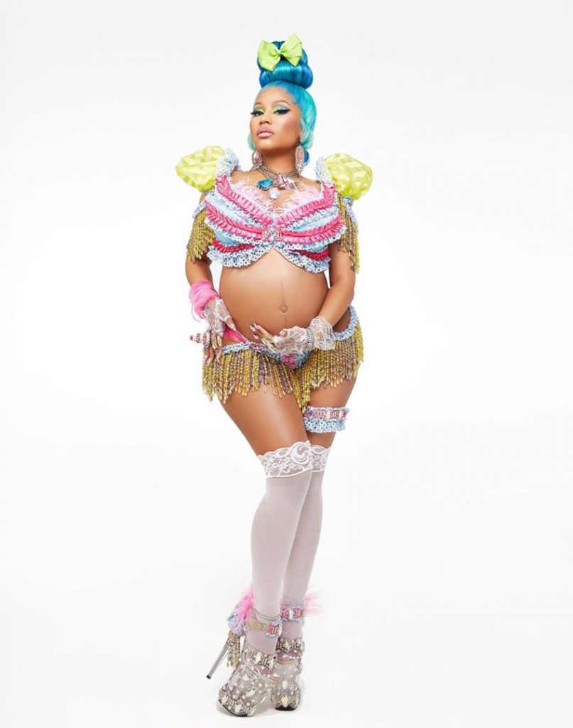 Nicki Minaj is standing in platform heels with turquoise hair and a bikini with knee socks cradling her pregnant belly