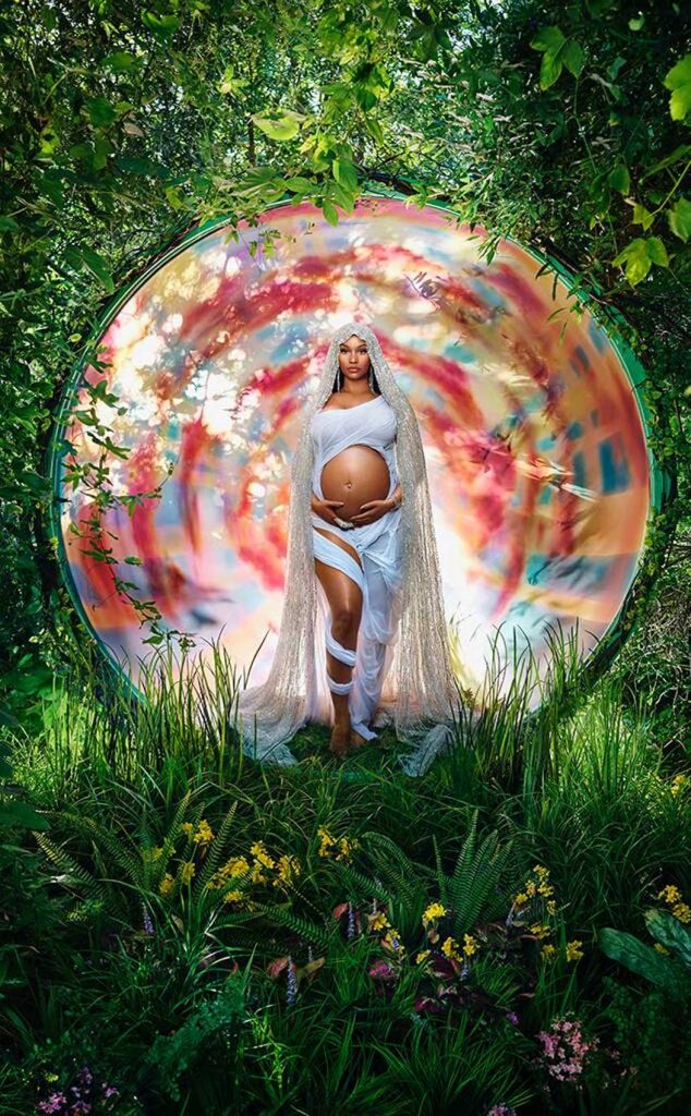 nicki minaj is in a colorful circle standing in a wooded area cradling her pregnant belly dressed in white