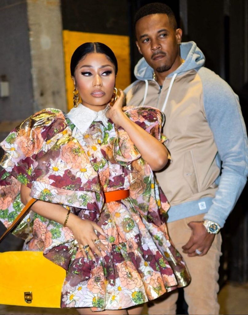 nicki minaj standing with her husband. she is wearing a colorful dress and he is wearing a blue and tan hoodie and khaki pants